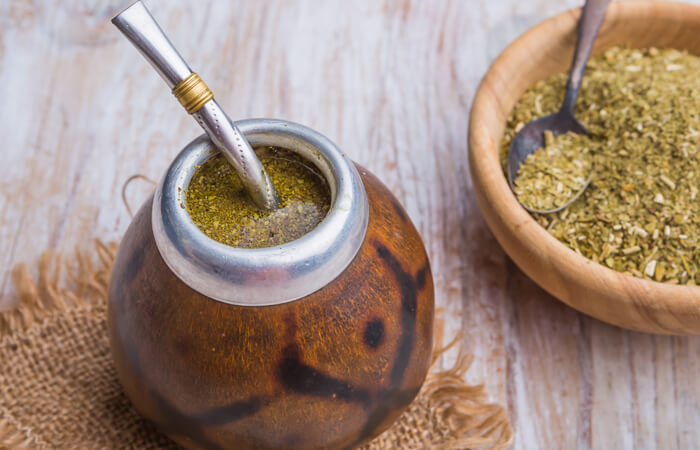 Yerba mate tea in a traditional gourd with a bombilla, accompanied by a wooden bowl of dried yerba mate leaves on a rustic wooden surface.
