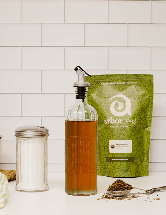 glass bottle of mint simple syrup next to sugar dispenser and package of organic peppermint