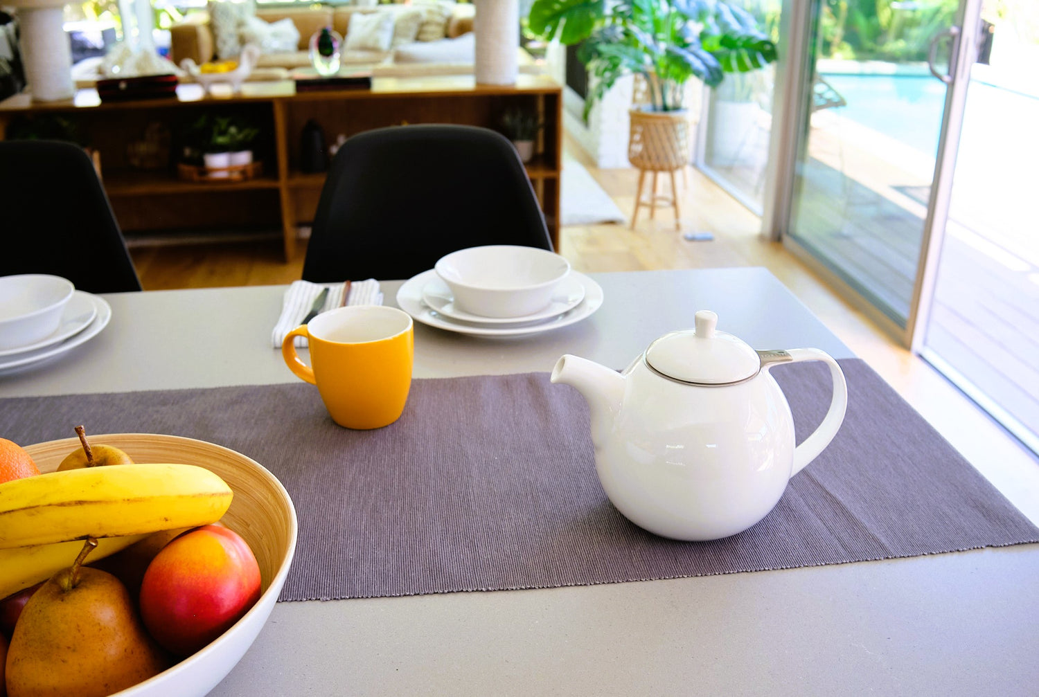Dining table set with a white Curve teapot, cups, and fruit in a bright room.