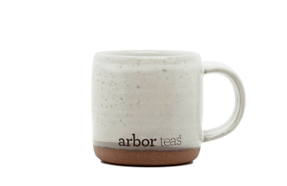 White ceramic mug with speckled pattern and 'arbor teas' branding on a black background